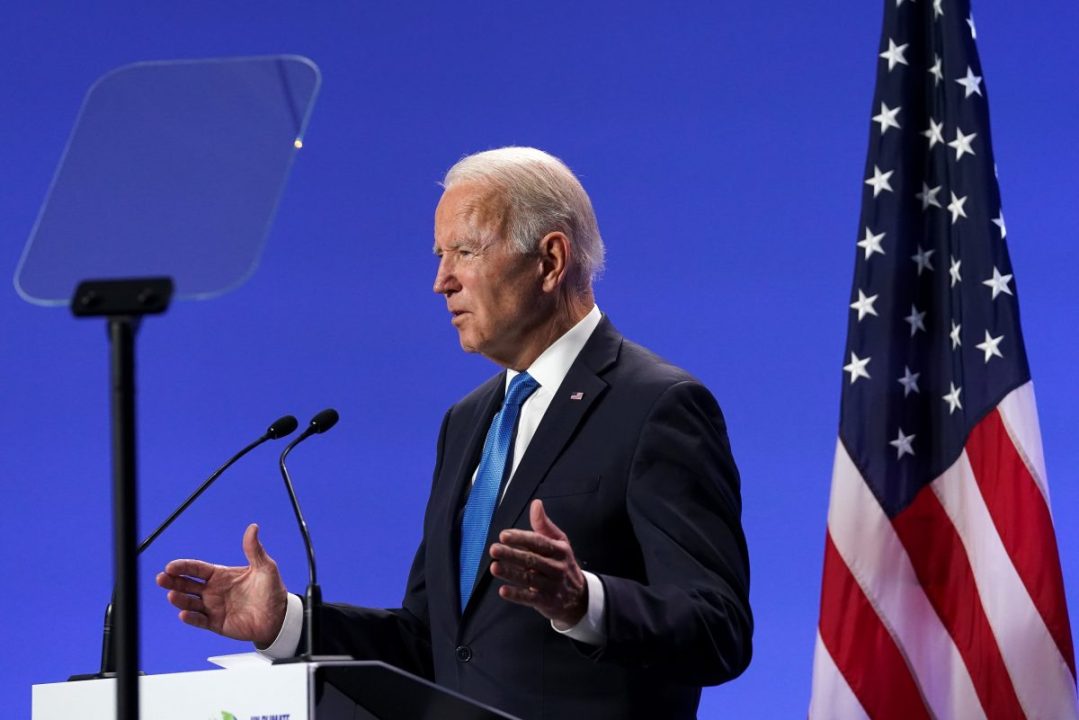 Joe Biden em palanque com fundo azul e bandeira dos EUA à direita
