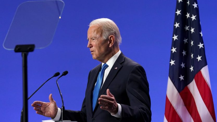 Joe Biden em palanque com fundo azul e bandeira dos EUA à direita