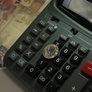 Foto de uma moeda de um real repousando sobre uma calculadora com notas de dinheiro ao fundo