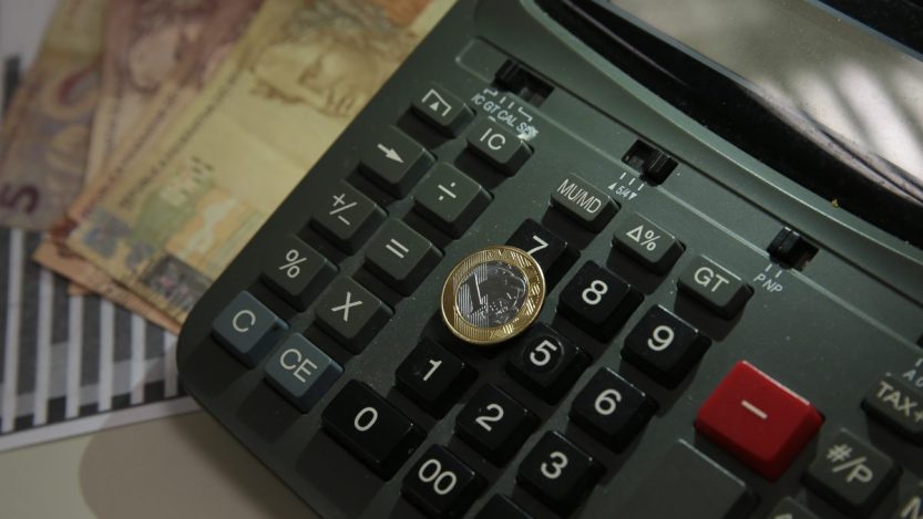 Foto de uma moeda de um real repousando sobre uma calculadora com notas de dinheiro ao fundo
