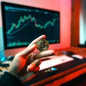 Foto de uma mão segurando uma moeda com o símbolo do bitcoin. Ao fundo uma tela de computador com gráficos.