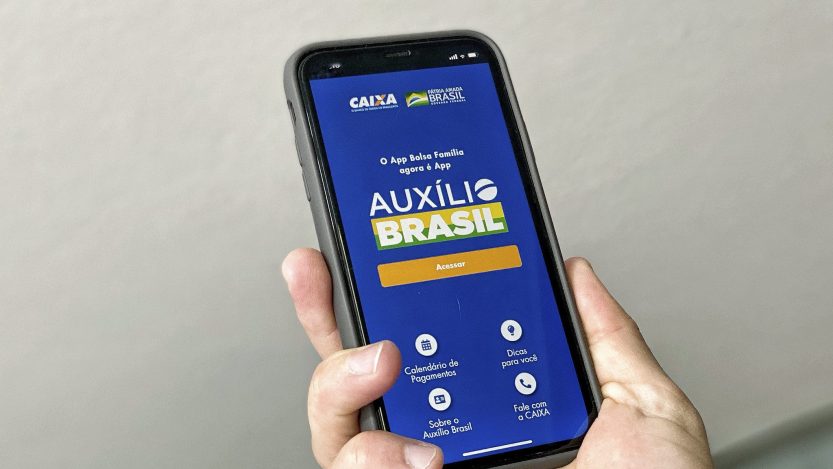 Mão segura celular em que tela mostra o aplicativo do Auxílio Brasil em fundo azul