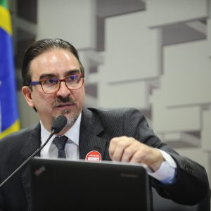 O economista Bernard Appy fala a um microfone com a bandeira do Brasil no fundo