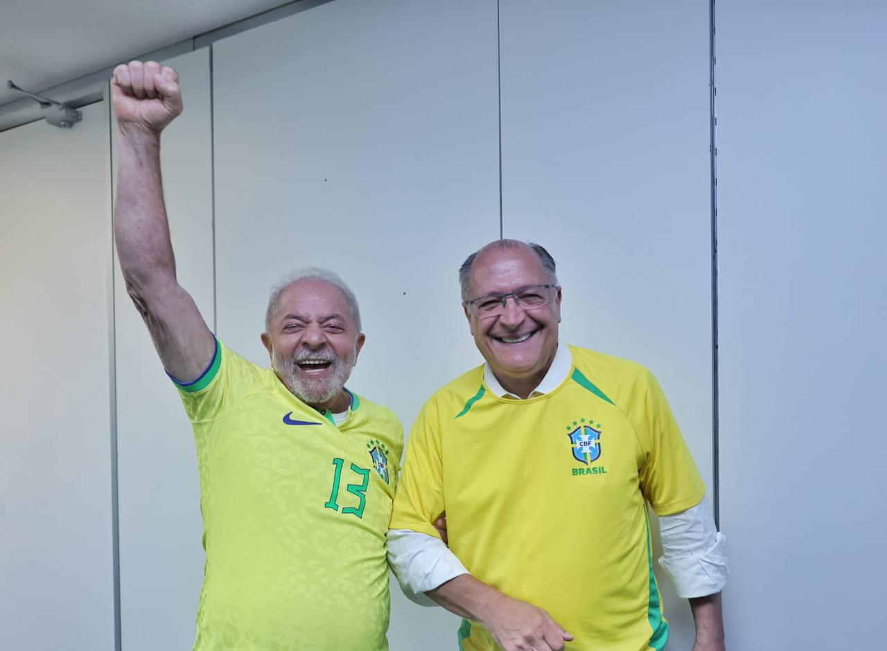 Foto de Luiz Inácio Lula da Silva e Geraldo Alckmin vestidos com a camisa amarela da seleção brasileira. Lula faz um gesto de levantar o braço em comemoração