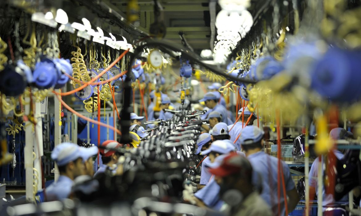 Foto de uma linha de produção industrial, onde diversas pessoas operam o mesmo tipo de maquinário