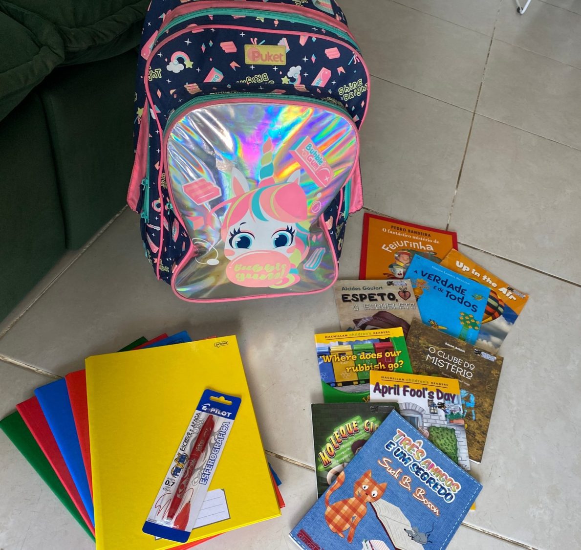 Imagem de materiais escolares: uma mochila, livros infantis, cadernos coloridos e uma caneta.