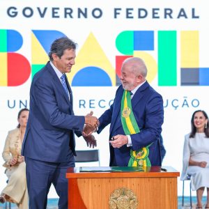 Foto de Fernando Haddad, ministro da Fazenda, cumprimentando Lula com a mão direita. Ao fundo, está o logo do governo federal com os dizeres: "Brasil. União e reconstrução"