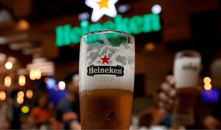 Foto de um copo de cerveja da Heineken com cerveja preenchendo a metade do copo com espuma no topo. Ao fundo está o logo da marca