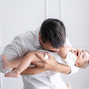 Homem segurando a filha pequena no colo. A menina está sorrindo. Ele está beijando a sua barriga