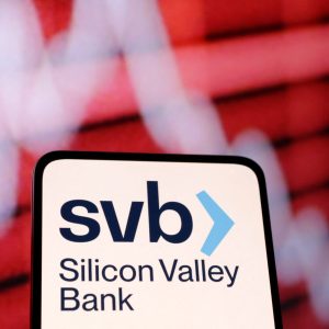 Foto de uma tela de celular com o logo do Banco do Vale do Silício (SVB, em inglês). Ao fundo, uma tela vermelha com um gráfico em queda.
