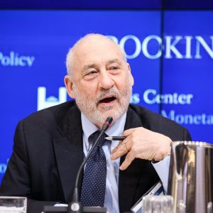 Foto de Joseph Stiglitz, Nobel em Economia. Ele é homem, tem barba e cabelo grisalho, veste um terno preto, camisa social azul-clara e gravata azul-escura. Ele também fala a um microfone