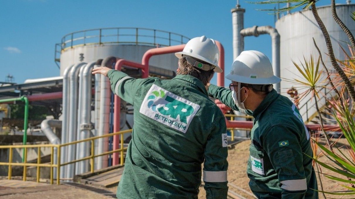 Foto de dois funcionários da 3R Petroleum (RRRP3) usando uniforme verde e capacete branco.