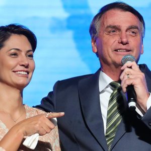 Foto de Jair Bolsonaro e Michele Bolsonaro, que está a seu lado esquerdo. Ela aponta com o dedo enquanto ele segura um microfone