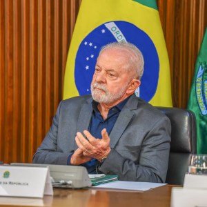 Foto do presidente Luiz Inácio Lula da Silva, vestindo terno cinza e camisa social azul marinha, de mãos cruzadas e sentado em uma cadeira