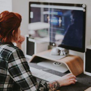 Foto de uma mulher ruiva e que usa camisa xadrez em frente ao computador, trabalhando.