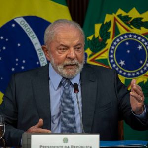 Foto de Luiz Inácio Lula da Silva, presidente da República. Ele usa um terno preto, gravata com estampas e camisa azul clara. Fala em um microfone com as bandeiras do Brasil e do governo federal ao fundo
