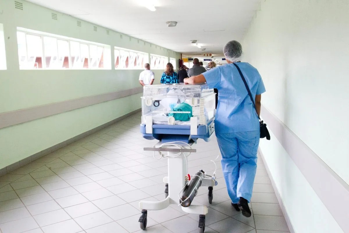 Foto de um enfermeiro em um hospital, carregando um leito. Ele veste um jaleco azul e usa toca. Foto ilustra matéria sobre efeitos do piso de enfermagem em ações de empresas na Bolsa