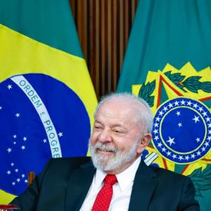 Foto de Luiz Inácio Lula da Silva, presidente da República, sorrindo. Ele veste um terno preto, camisa branca e grava vemelha. Ao fundo, uma bandeira do Brasil e uma bandeira com o brasão da Presidência da República