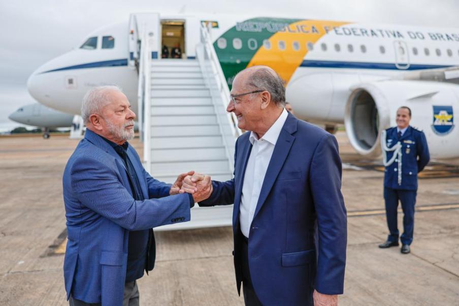 Foto de Luiz Inácio Lula da Silva, presidente do Brasil, cumprimentando Geraldo Alckmin, vice-presidente do Brasil. Eles se cumprimentam perante o avião presidencial com faixas verdes e amarelas. A escada de embarque está posicionada