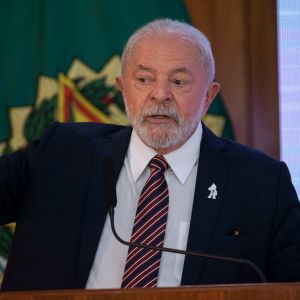 Foto de Luiz Inácio Lula da Silva, presidente da República. Ele fala a um microfone, gesticula com a mão esquerda e veste um terno preto, camisa branca e gravata listrada vermelha e preta.