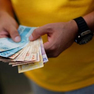 Mãos de homem conta dinheiro renegociado pelo Desenrola Brasil; ele veste uma camiseta amarela e tem um relógio preto em seu pulso esquerdo