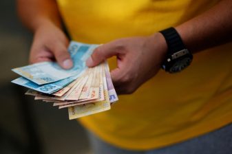 Mãos de homem conta dinheiro renegociado pelo Desenrola Brasil; ele veste uma camiseta amarela e tem um relógio preto em seu pulso esquerdo