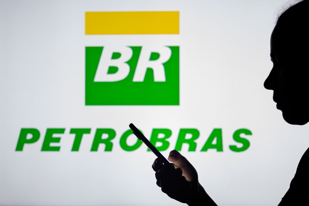Foto do logo da Petrobras com uma pessoa usando o celular em primeiro plano. A matéria descreve como reinvestir dividendos das ações (PETR3;PETR4) dobrou retorno no 1º semestre