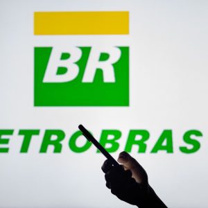 Foto do logo da Petrobras com uma pessoa usando o celular em primeiro plano. A matéria descreve como reinvestir dividendos das ações (PETR3;PETR4) dobrou retorno no 1º semestre