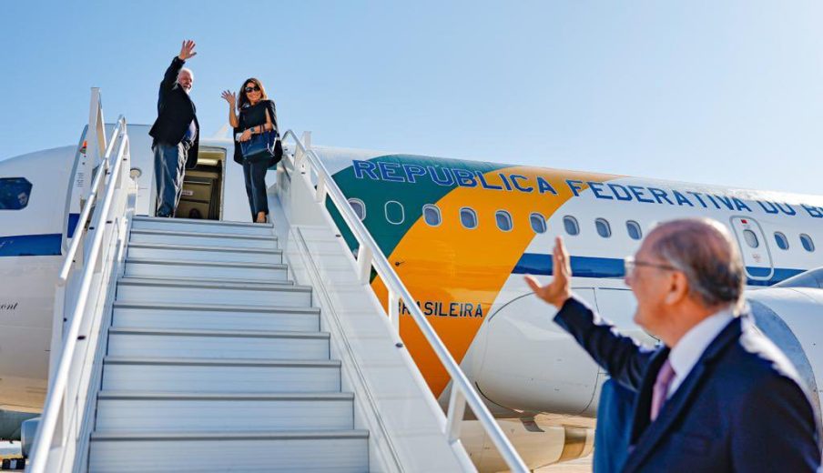 Foto do presidente Luiz Inácio Lula da Silva e da primeira-dama Janja Lula da Silva acenando, do topo de uma escada para a aeronave presidencial.