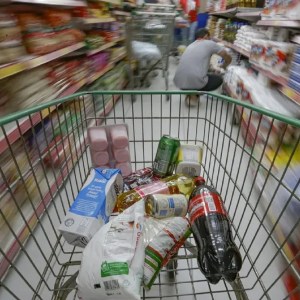 Foto de um carrinho de supermercado com produtos. Ao fundo, um consumidor procura produtos em uma prateleira de um supermercado