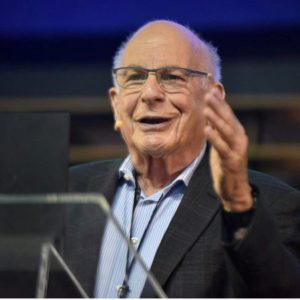 Foto de Daniel Kahneman, psicólogo e economista vencedor do Nobel em Economia. Na imagem, ele está discursando e de braços erguidos.