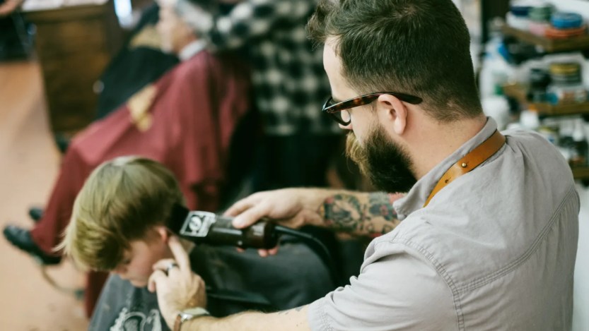 Foto de um cabelereiro cortando cabelo de um cliente. Ele é branco, usa óculos e usa uma máquina para cortar cabelo
