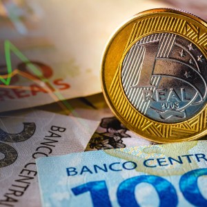 Foto de uma moeda de um real sobre uma nota de cem reais. Matéria explica sobre aumento de rating do Brasil pela agência Standard&Poors