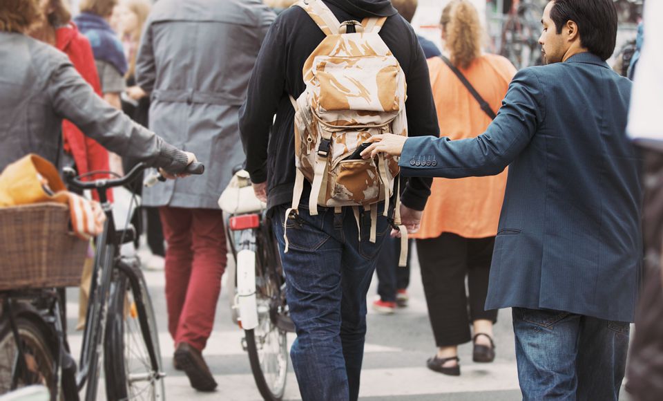 A imagem mostra algumas pessoas de costas andando. Uma delas está com uma mochila. Ao lado há um homem de terno roubando um item de mochila.