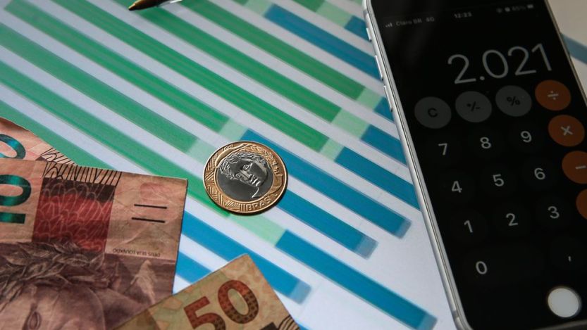 Foto mostra notas de R$ 10 e R$ 50, moeda de R$1 e uma calculadora em um aparelho celular