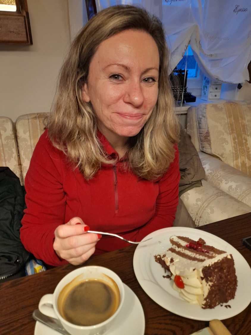 Fernanda Bottoni é loira, tem cabelos na altura dos ombros, usa uma camisa vermelha, come um grande pedaço de bolo de creme e toma um café