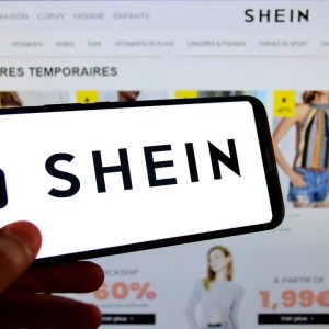 A imagem mostra uma mão segurando um celular escrito "Shein" na tela. Ao fundo está uma página de e-commerce com anúncios de roupas.