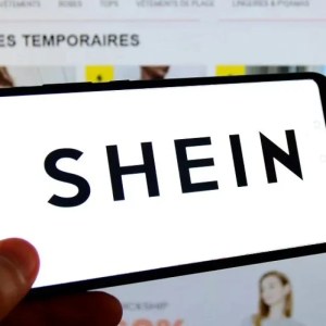 A imagem mostra uma mão segurando um celular escrito "Shein" na tela. Ao fundo está uma página de e-commerce com anúncios de roupas.