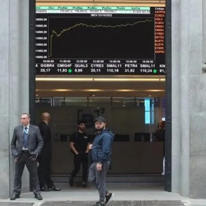 A imagem mostra a fachada da B3, bolsa de valores brasileira, com algumas pessoas passando na frente.