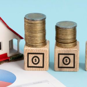 Foto para a matéria sobre quanto rendem R$ 100 em fundos imobiliários em que aparece uma maquete de uma casa em cima de um papel com gráficos e, ao lado, três blocos com pilhas de moedas por cima.