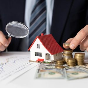 Foto de um homem segurando uma lupa, empilhando moedas com uma miniatura de casa em cima de papeis de investimentos. Imagem para a matéria sobre quanto rende R$ 1 milhão em fundos imobiliários.