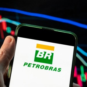 Foto do logo da Petrobras (PETR3;PETR4) em uma tela de celular com fundo branco e gráficos de vela ao fundo.