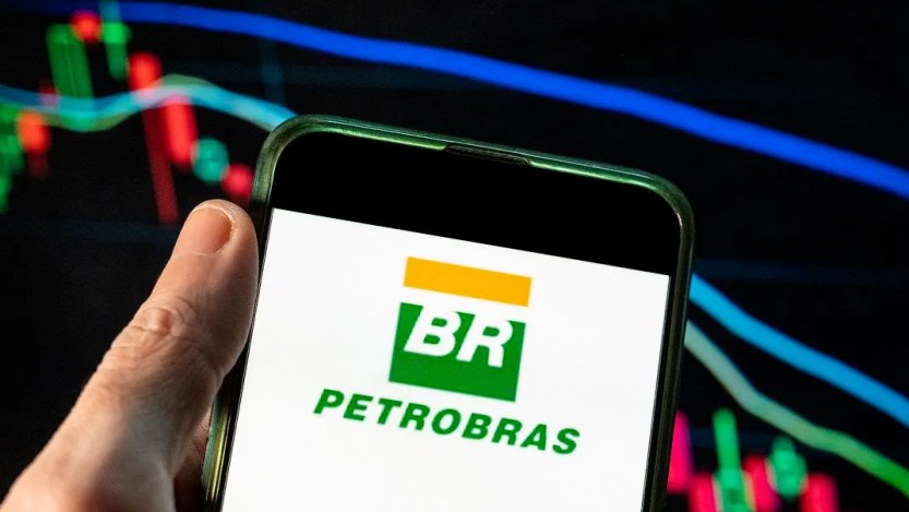 Foto do logo da Petrobras (PETR3;PETR4) em uma tela de celular com fundo branco e gráficos de vela ao fundo.