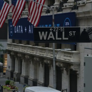 Imagem da fachada da Bolsa de Nova York, em Wall Street, com algumas bandeiras dos Estados Unidos.