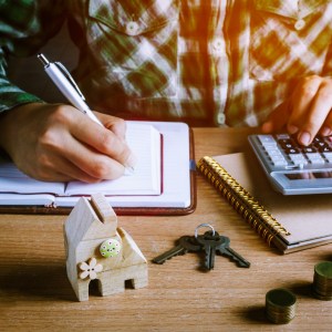 Imagem para o texto sobre amortizar financiamento em que aparece um homem fazendo cálculos com uma maquete de uma casa em cima da mesa, um molho de chaves e três pilhas de moedas.