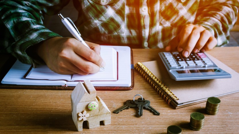 Imagem para o texto sobre amortizar financiamento em que aparece um homem fazendo cálculos com uma maquete de uma casa em cima da mesa, um molho de chaves e três pilhas de moedas.