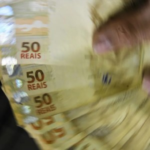 Foto de uma pessoa segurando notas de R$ 50. A matéria explica como investir para criar uma carteira de renda mensal