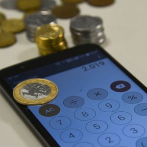 Imagem mostra calculadora de celular e moedas de real para ilustrar reportagem sobre quanto rende R$ 1 milhão no Tesouro Selic?