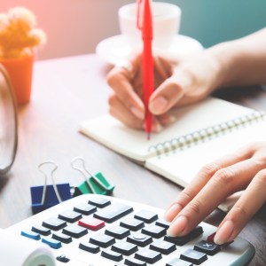 Imagem de uma mão fazendo cálculos em uma calculadora e a outra mão escrevendo com uma caneta vermelha em um caderno para a matéria sobre viver de renda com 100 mil reais.