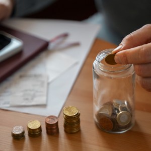 Imagem de uma mão colocando moedas dentro de um pote ao lado de várias pilhas de moeda e uma calculadora. Imagem para a matéria sobre viver de renda com 500 mil
